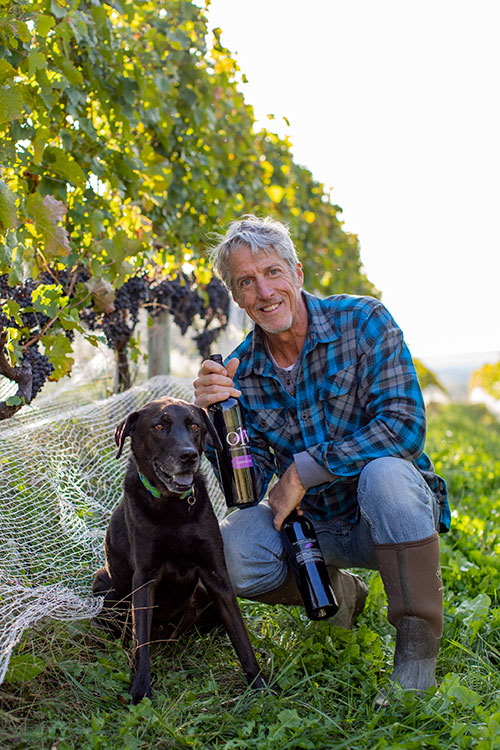 Bob Rupy holding Wine bottle with Becca Pup by vines to represent the Virginia Wine Club